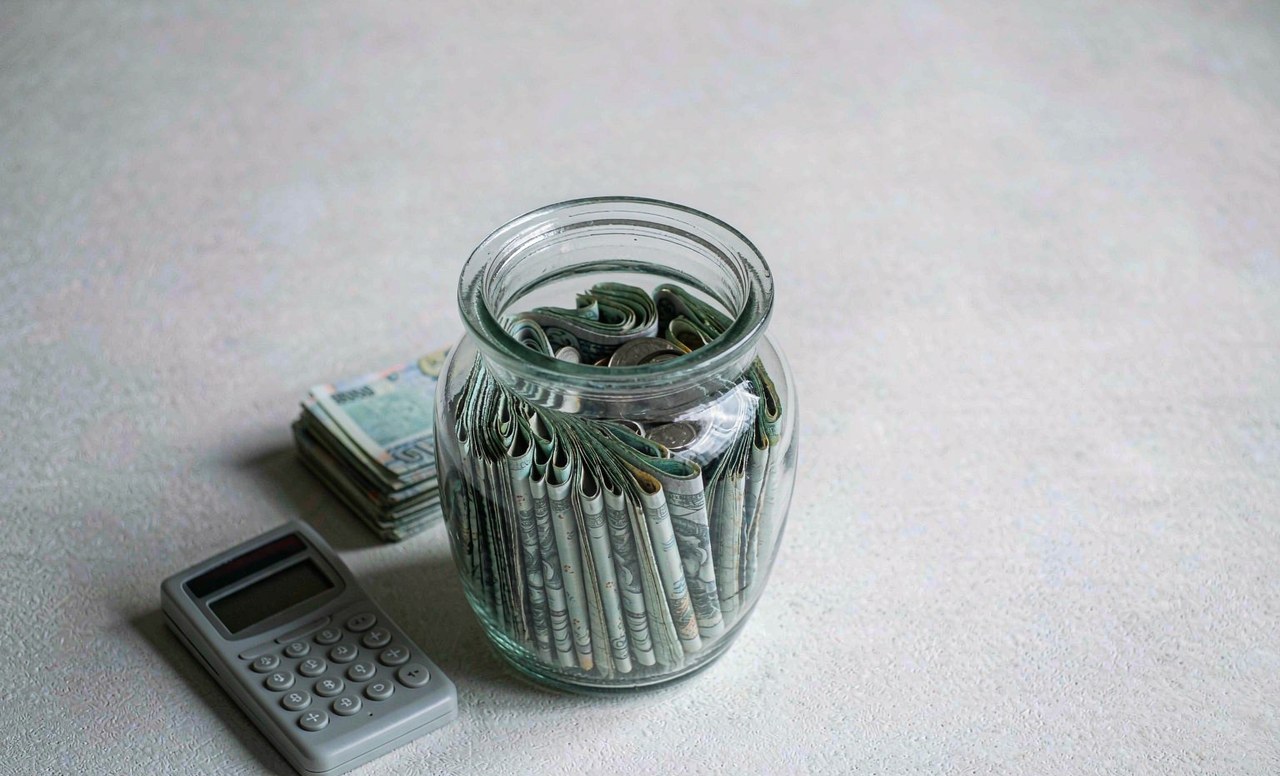 A savings jar and a stack of banknotes on a simple desk with a calculator