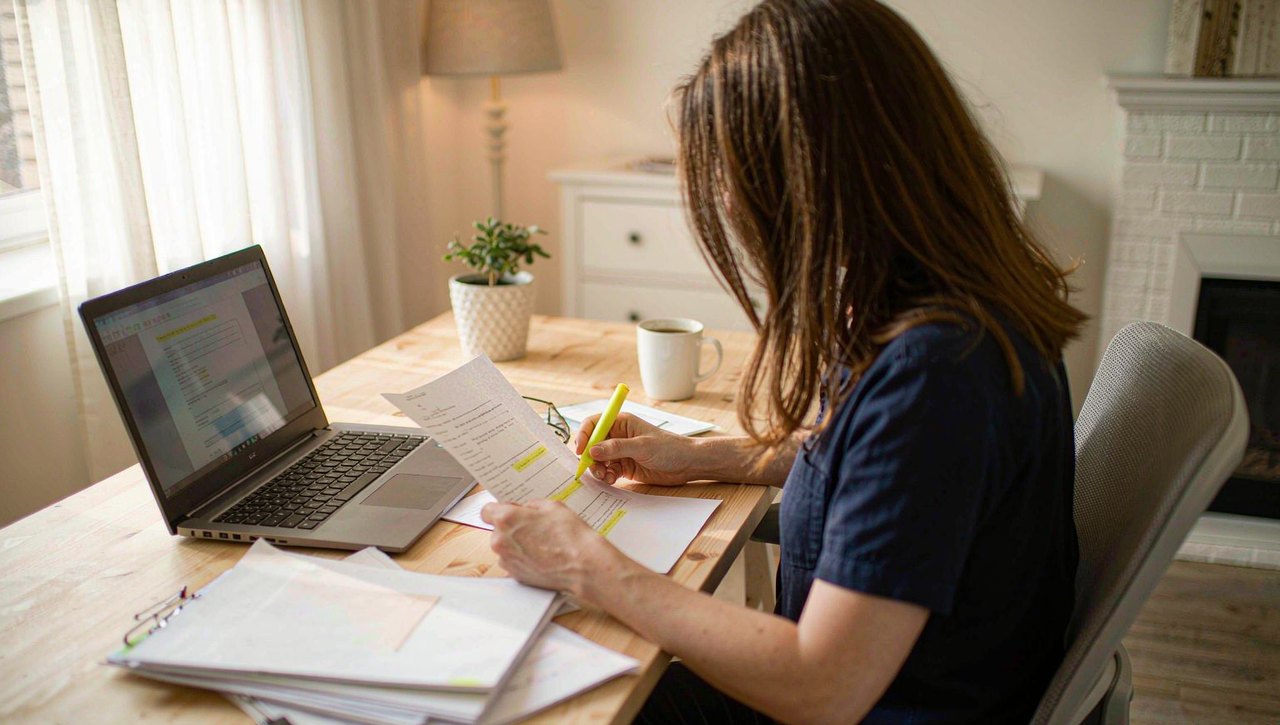 A person at a home desk reviewing printed pages with a highlighter beside an open laptop, glasses and a coffee mug nearby, warm daylight